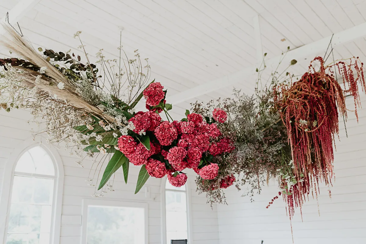 An elaborate hanging floral installation with vibrant pink flowers and dried grasses in a white-walled room.